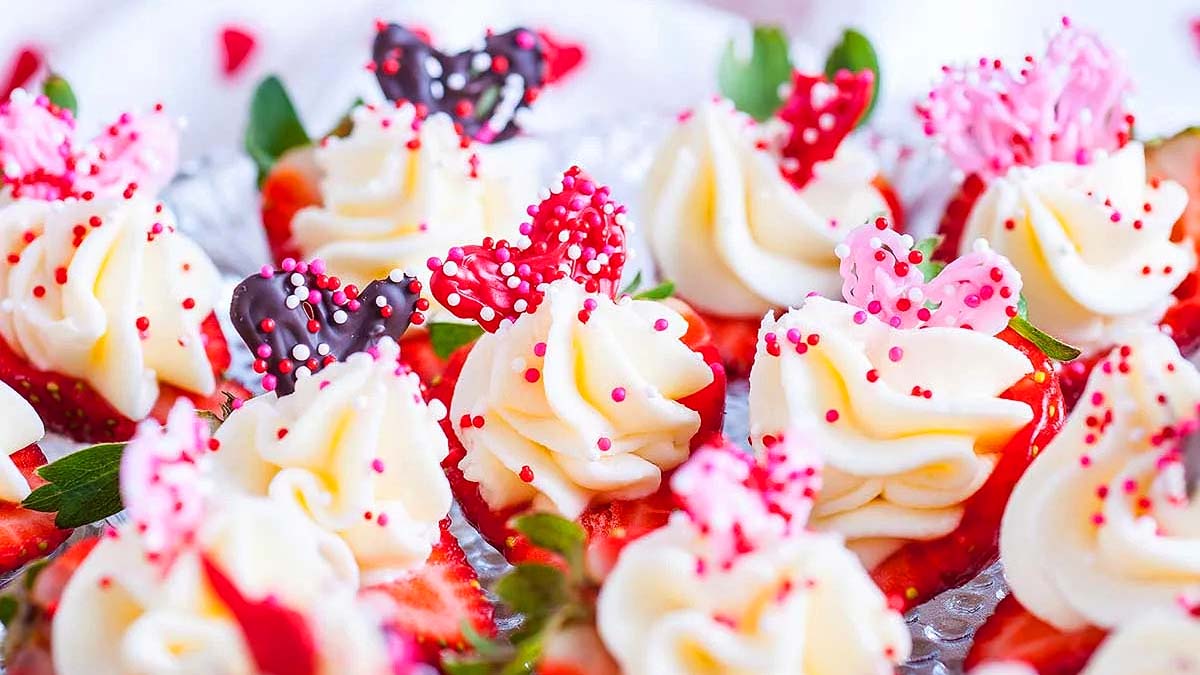 Close-up of desserts topped with swirls of white frosting, red and pink heart-shaped decorations, and colorful sprinkles on a bed of sliced strawberries.