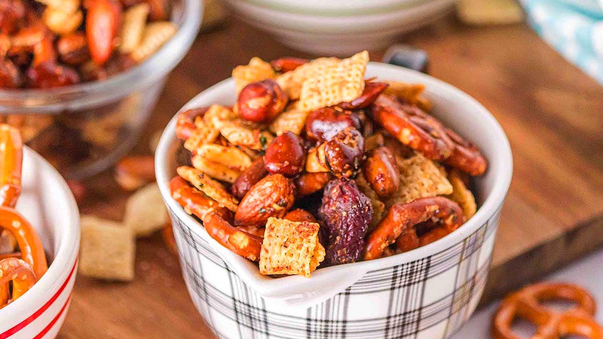A bowl of snack mix containing pretzels, cereal squares, nuts, and seasoned pieces sits on a wooden surface.