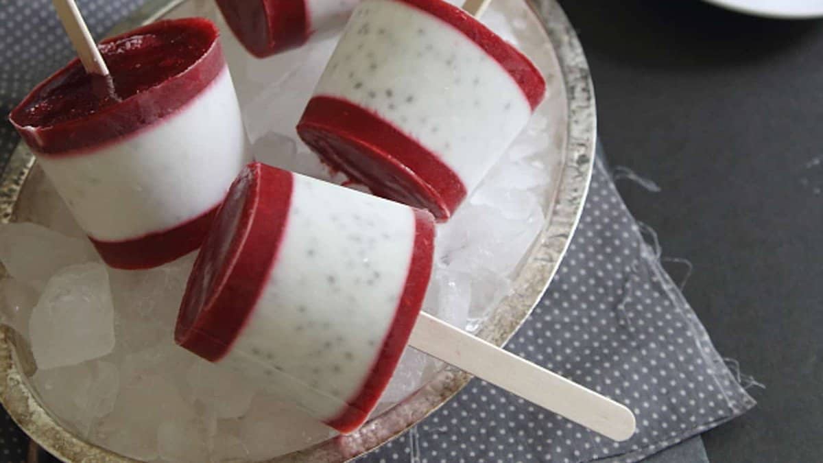 Four layered popsicles with white and red sections rest on a bed of ice in a silver tray, with one popsicle lying sideways and three standing upright.