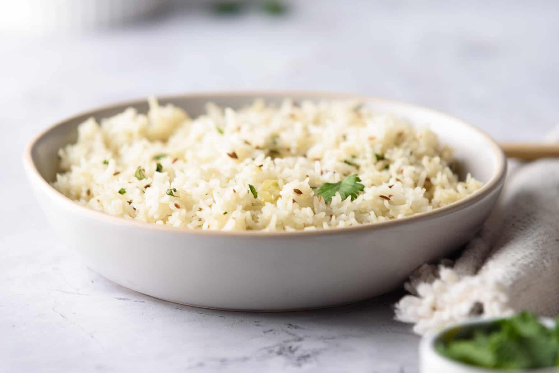 A shallow white bowl filled with cooked white rice garnished with herbs, placed on a light-colored surface next to a beige cloth.