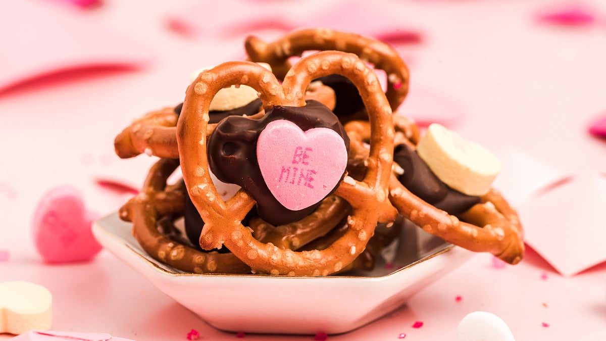 A small dish with heart-shaped pretzels topped with chocolate and pink candy hearts that say Be Mine, surrounded by more candy hearts on a pink surface.
