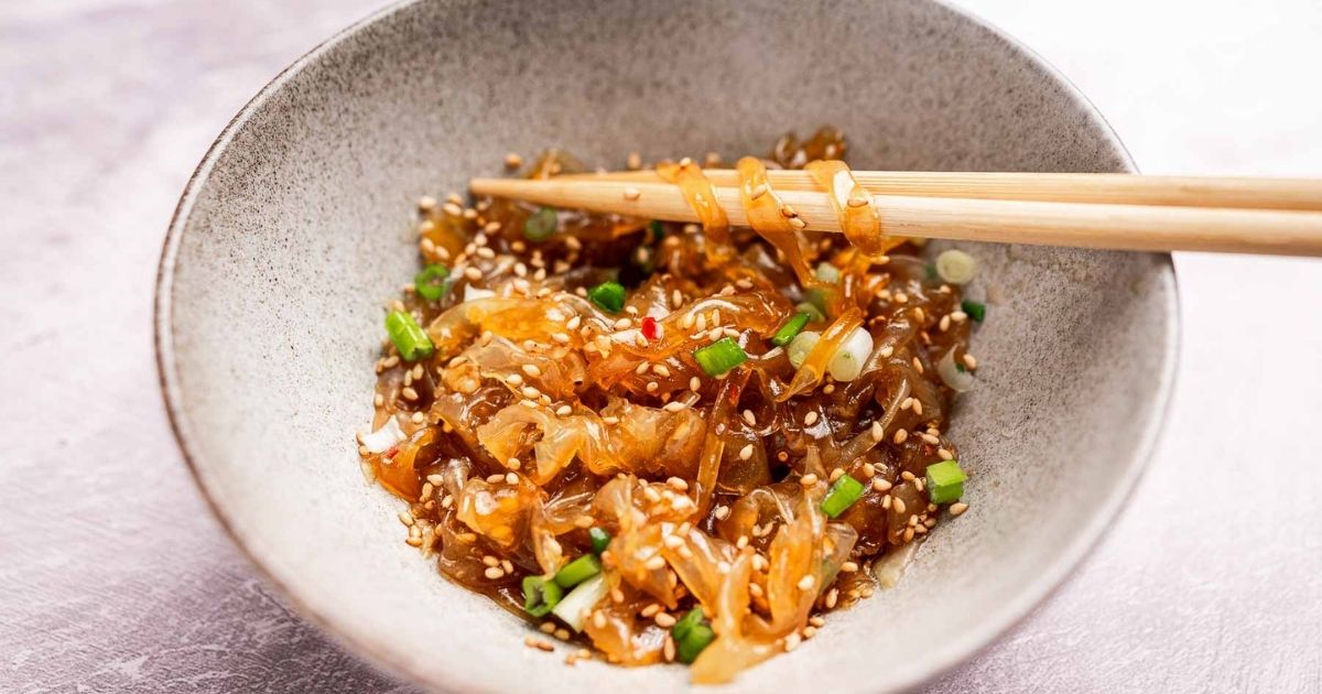 A bowl of seasoned jellyfish salad garnished with chopped green onions and sesame seeds, with chopsticks picking up a portion.