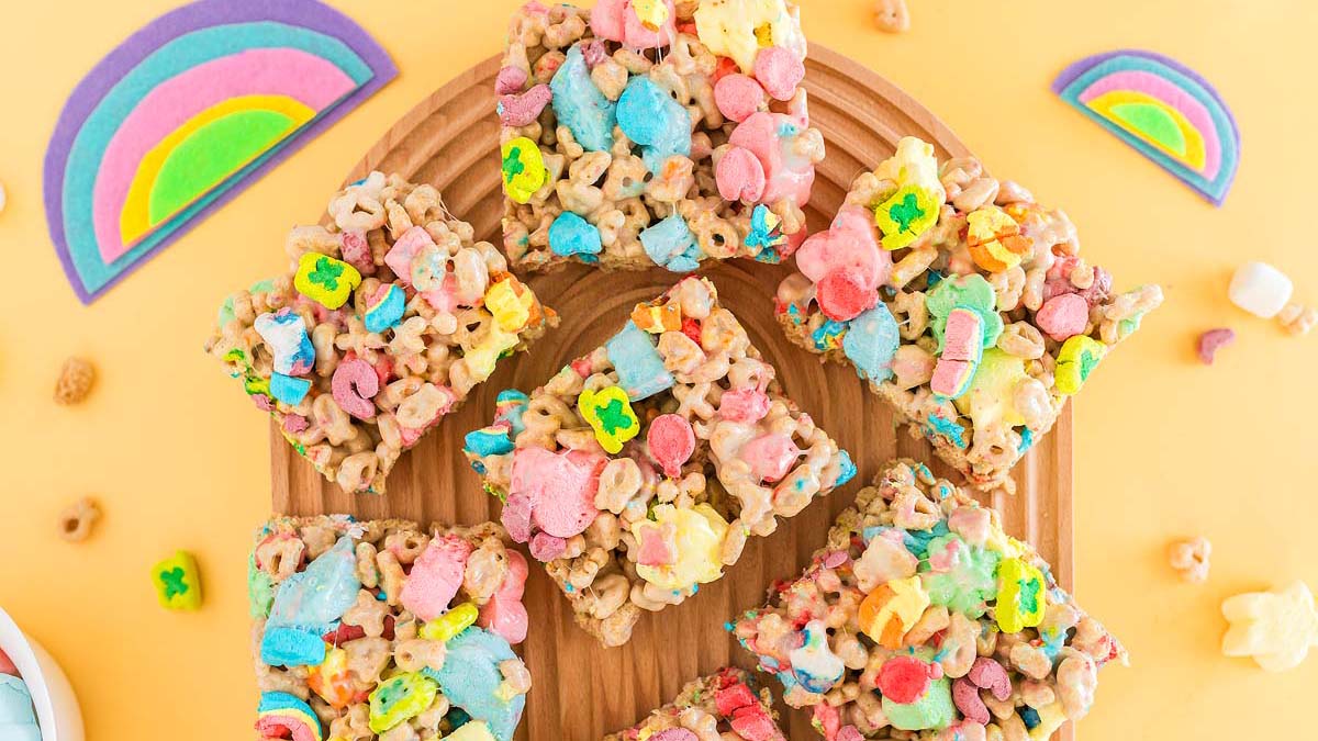 Six colorful cereal marshmallow bars are arranged on a wooden platter, surrounded by rainbow decorations on a yellow background.