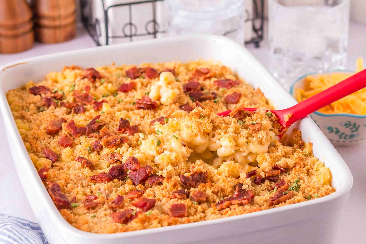 A baking dish of macaroni and cheese topped with breadcrumbs and bacon, with a serving spoon lifting a portion. Shredded cheese and glasses of water are in the background.