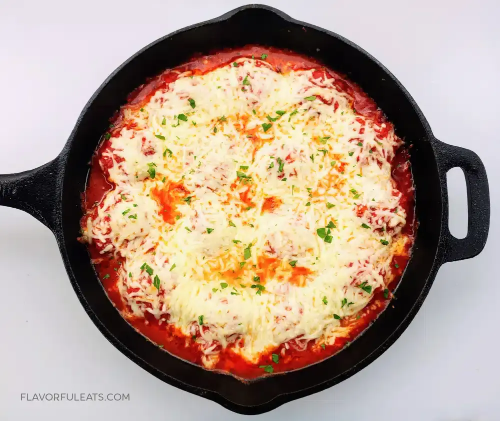 A skillet filled with baked cheesy marinara sauce, topped with melted cheese and garnished with chopped herbs.