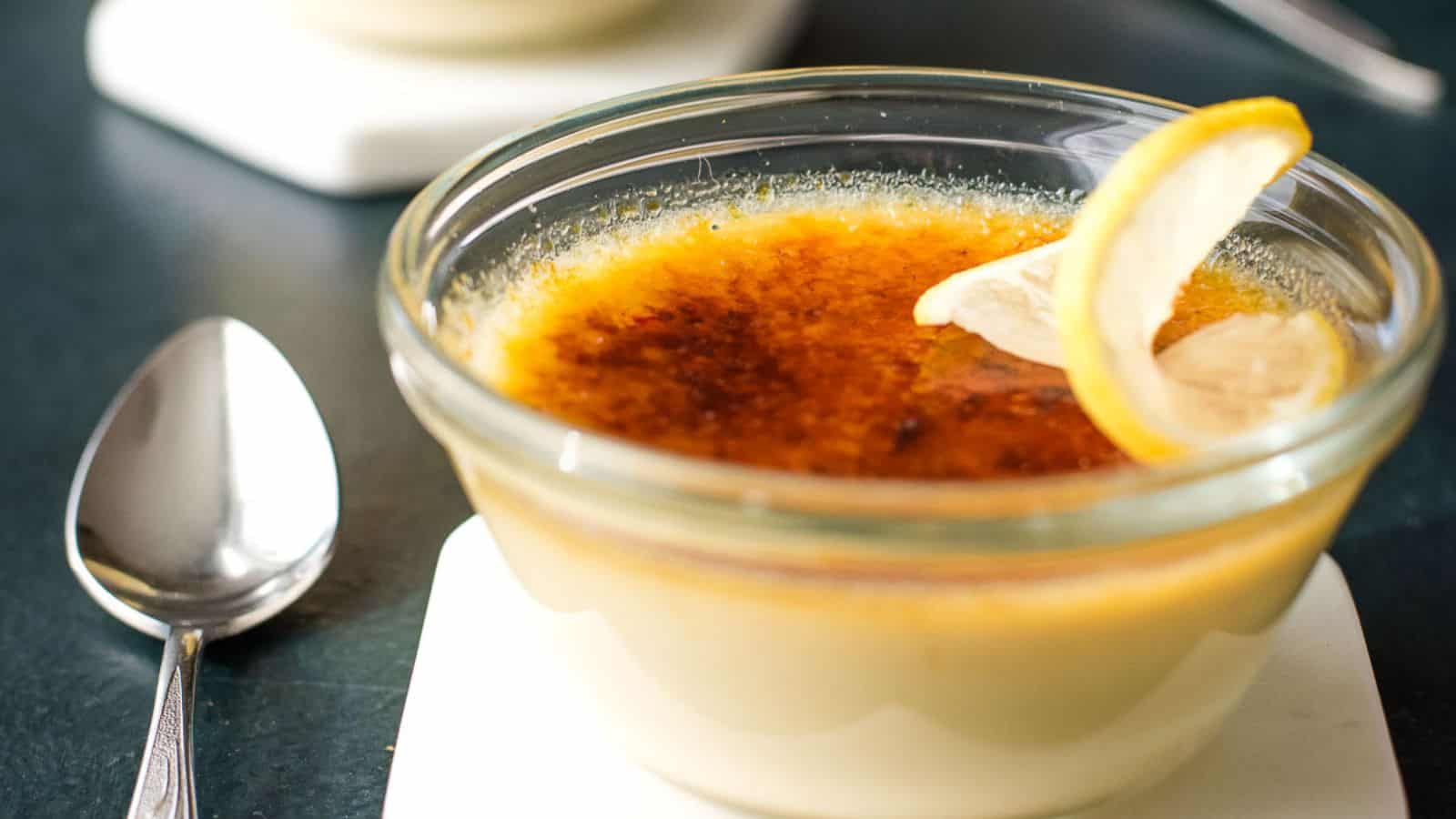 A glass bowl of crème brûlée garnished with two lemon slices, placed on a white coaster with a metal spoon nearby.