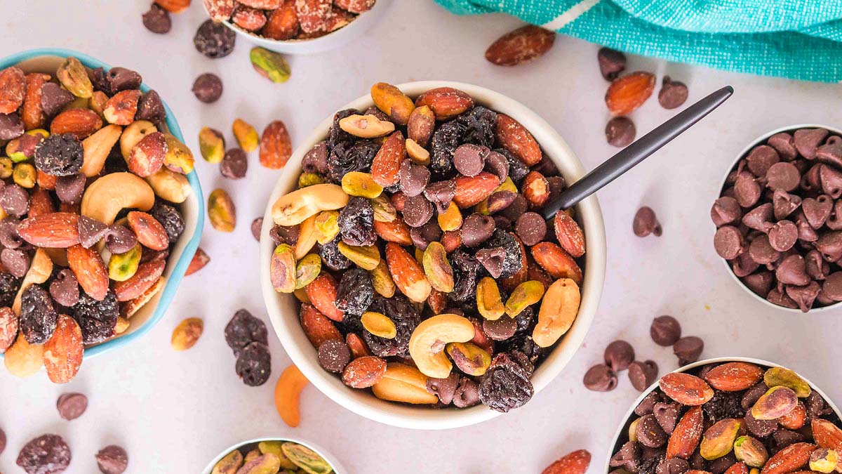A bowl of trail mix with nuts, dried fruit, and chocolate chips surrounded by small dishes of similar ingredients on a white surface.