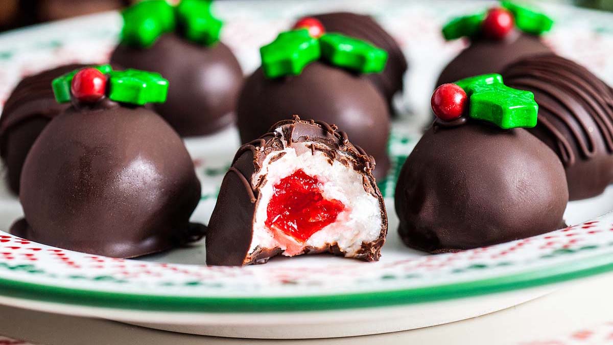Chocolate truffles decorated with green holly leaf and red berry candies, one cut open to reveal a white filling with a red center, arranged on a festive plate.