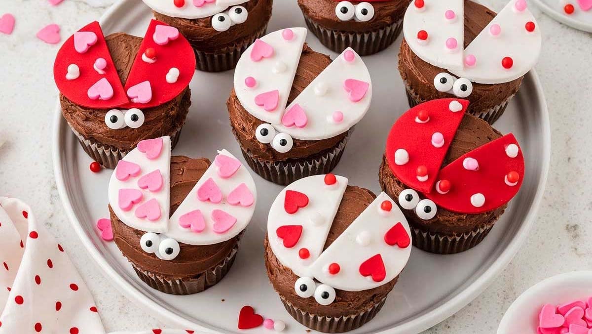 Chocolate cupcakes decorated with fondant to look like ladybugs, featuring red, pink, and white wings with heart and dot designs, and candy eyes, arranged on a round plate.
