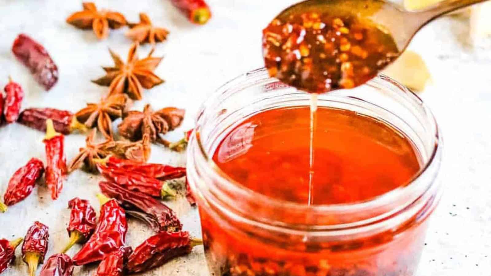 A jar of chili oil with visible chili flakes, a spoon dripping oil, dried red chilies, and star anise on a white surface.
