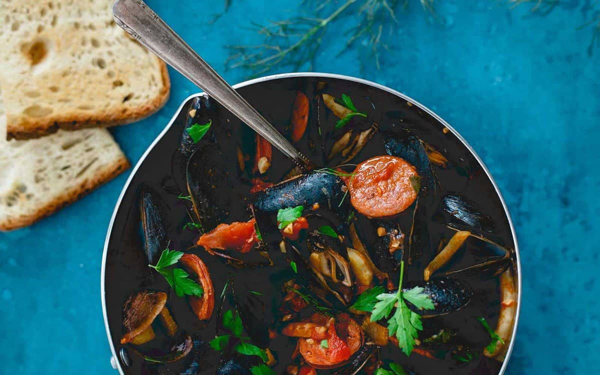 A bowl of mussel stew with tomatoes and herbs, placed on a blue surface next to slices of bread.