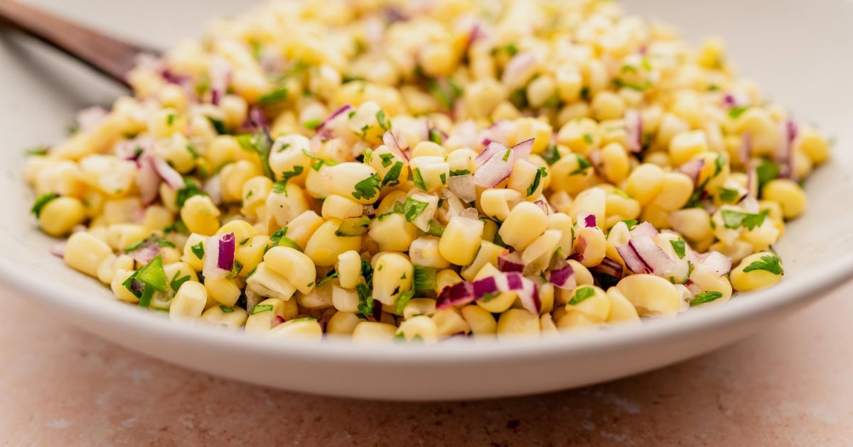 A close-up of a bowl filled with corn salad, featuring yellow corn kernels, chopped red onion, and green herbs.