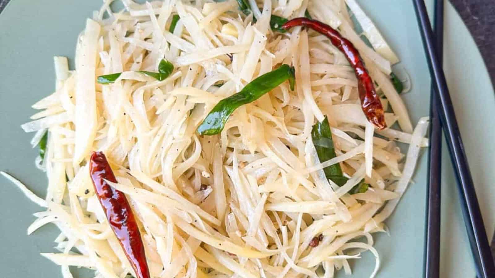 A plate of stir-fried shredded potatoes garnished with dried red chilies and green peppers, with chopsticks placed on the side.