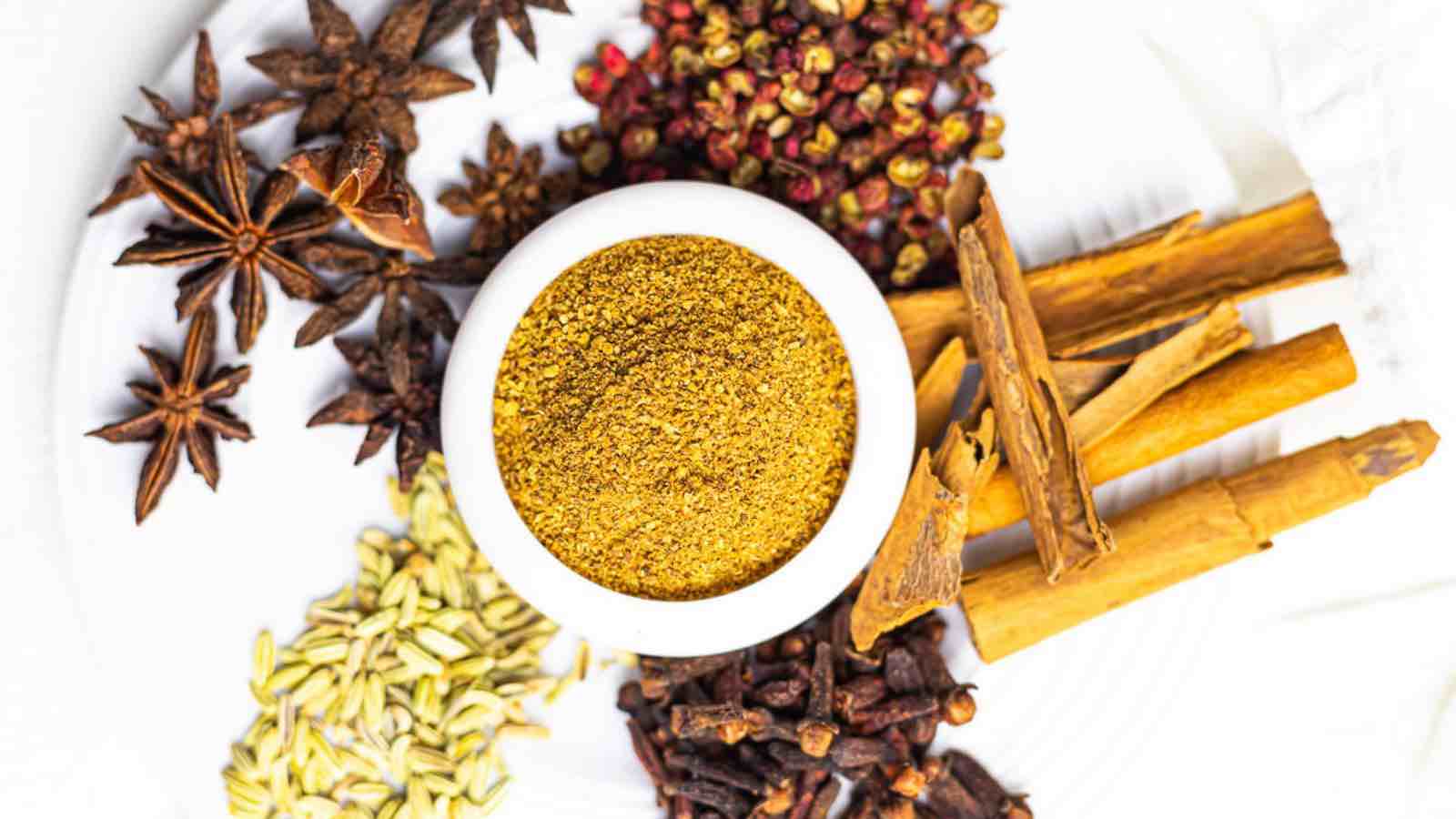 A bowl of ground spice mix is surrounded by whole spices including star anise, fennel seeds, cinnamon sticks, cloves, and Sichuan peppercorns on a white surface.