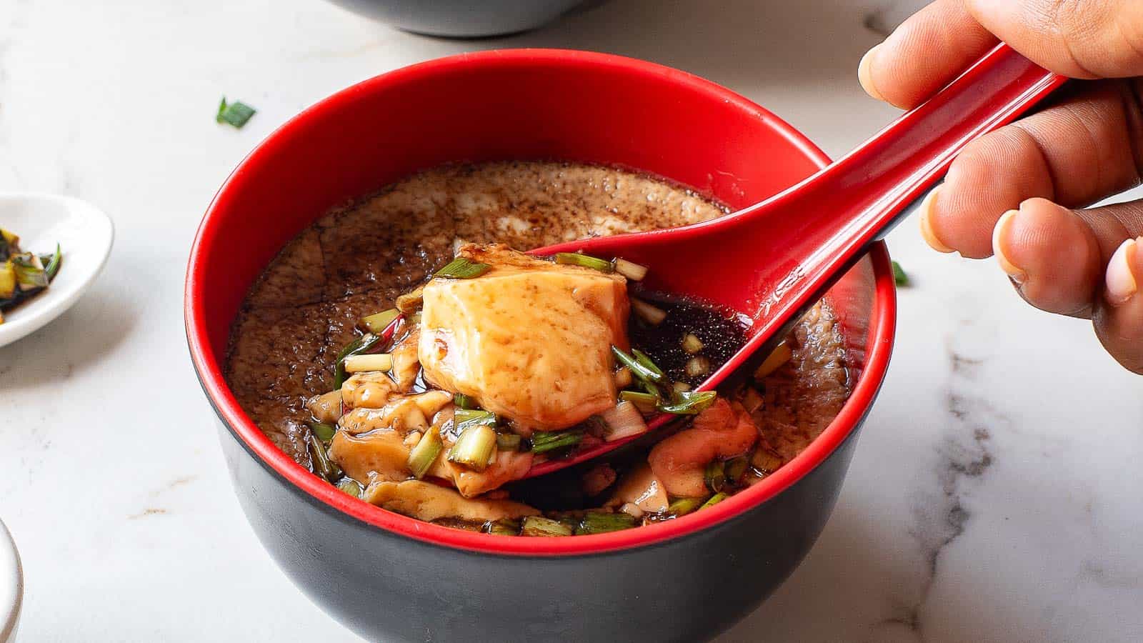 A hand holds a red spoon lifting a portion of savory steamed egg custard topped with sauce and chopped green onions from a red and black bowl.