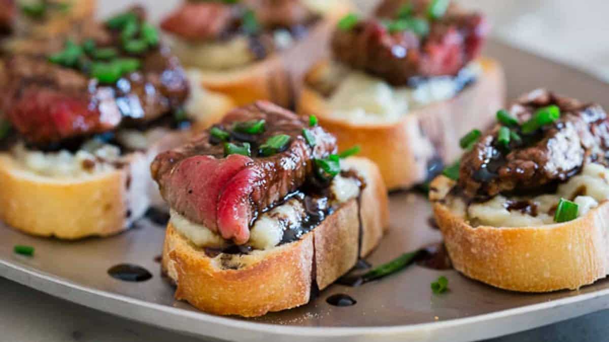 Slices of toasted baguette topped with blue cheese, steak, balsamic glaze, and chopped chives are arranged on a platter.