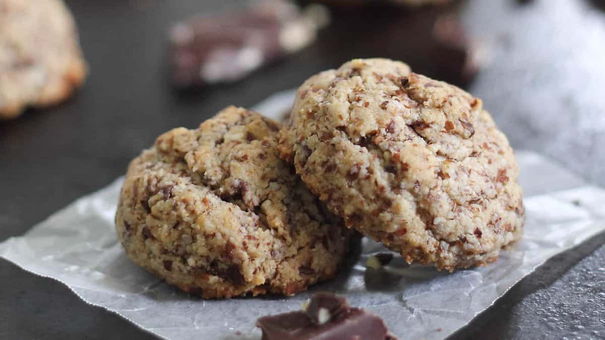 Two chocolate chip cookies rest on a piece of parchment paper, with pieces of chocolate scattered around on a dark surface.