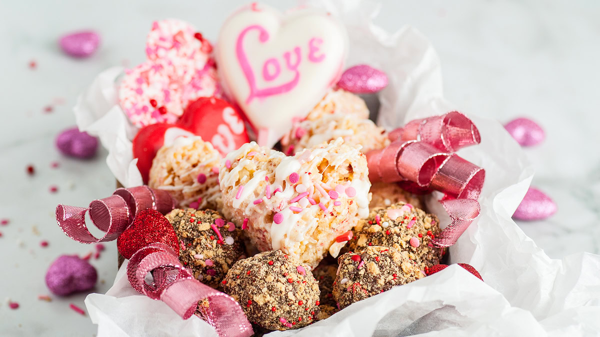 A box filled with heart-shaped and round treats, some decorated with pink and white icing, sprinkles, and ribbons, with a large cookie featuring the word Love.