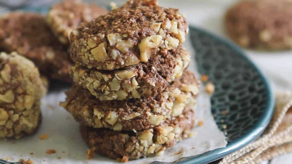 A stack of four chocolate cookies coated with chopped nuts sits on a blue plate, with more cookies in the background.