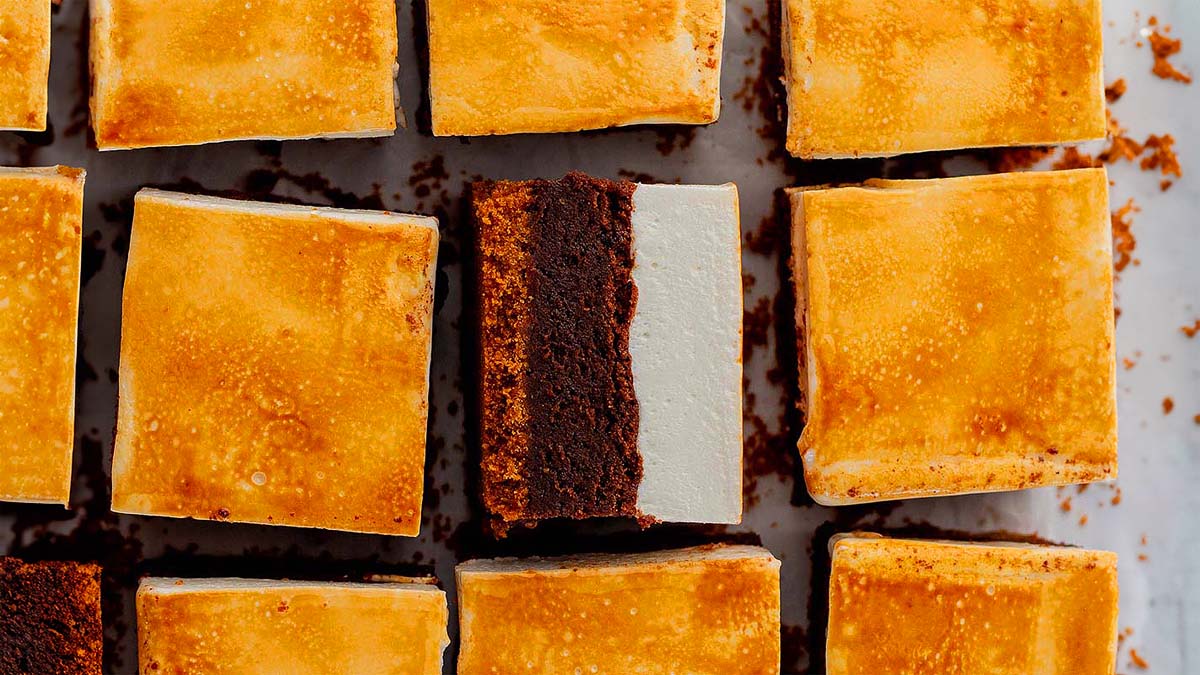Squares of dessert with a toasted golden top and a visible layer of chocolate brownie beneath arranged in rows on a white surface.