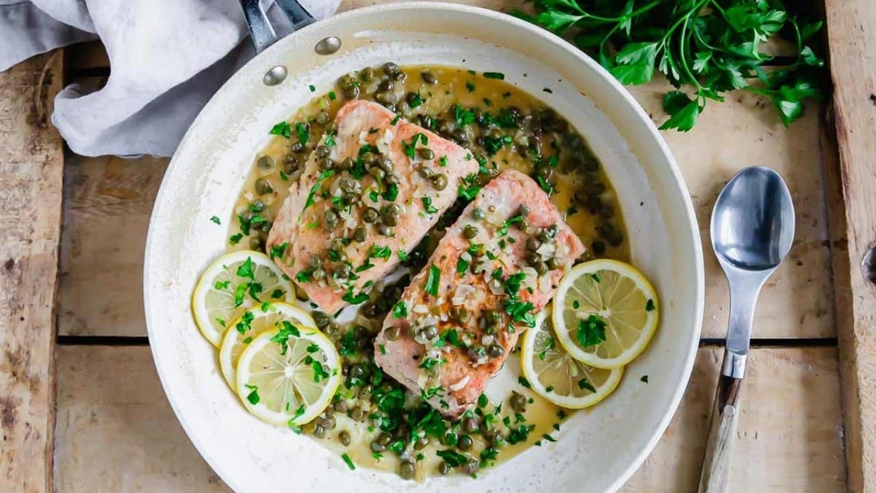 Two pieces of salmon in a white pan with capers, lemon slices, chopped parsley, and a creamy sauce, on a wooden table with a spoon and fresh parsley nearby.