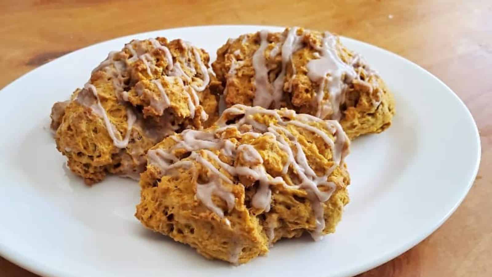 Three pumpkin scones with icing drizzle are arranged on a white plate set on a wooden surface.