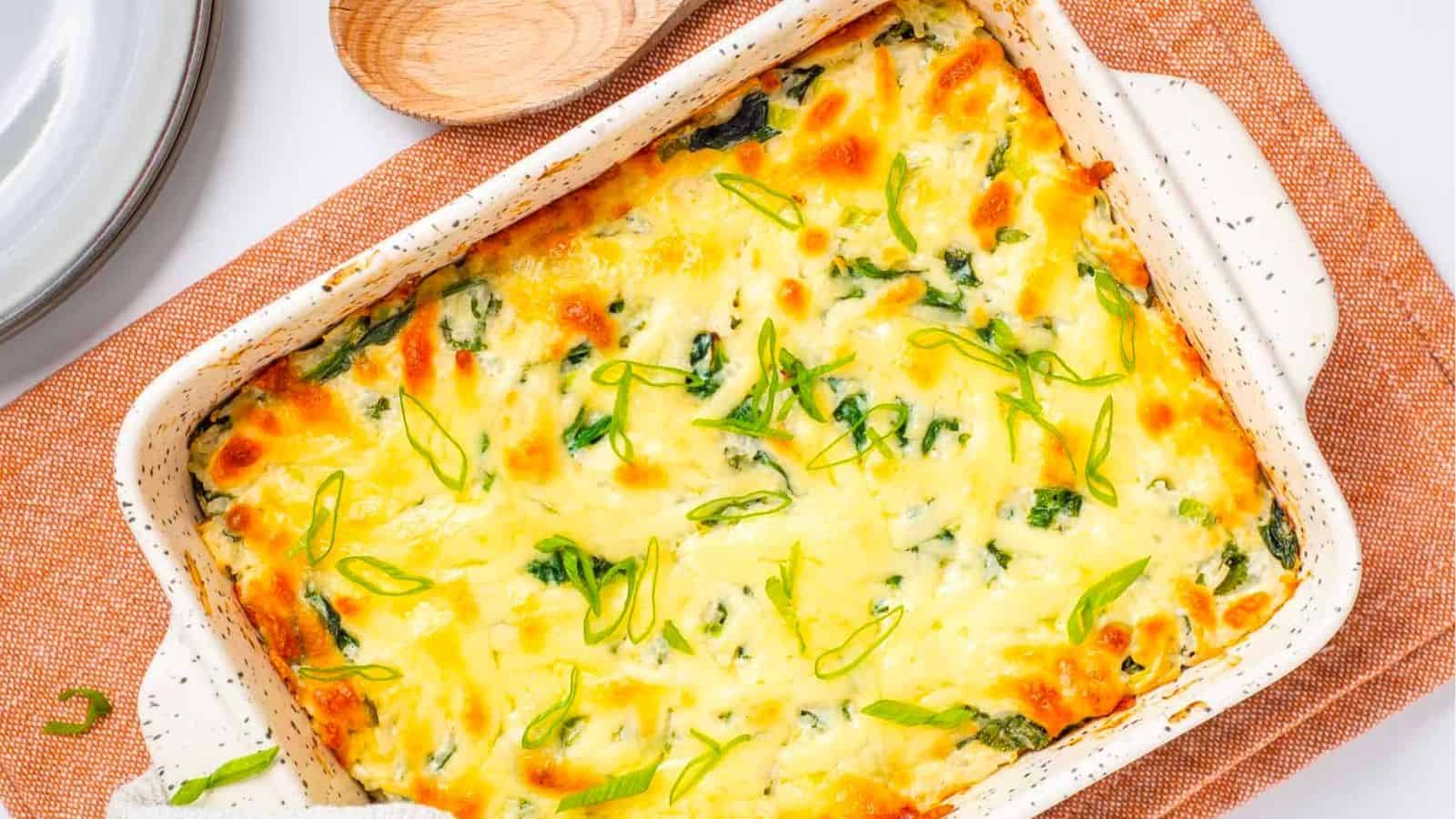 A baked casserole dish filled with a cheesy spinach casserole, topped with melted cheese and sliced green onions, on a brown cloth with a wooden spoon nearby.