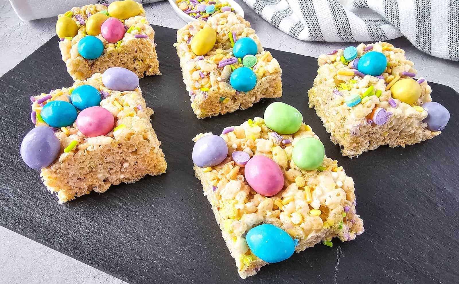 Five rice crispy treat squares topped with colorful candy eggs and sprinkles are arranged on a black slate serving board.