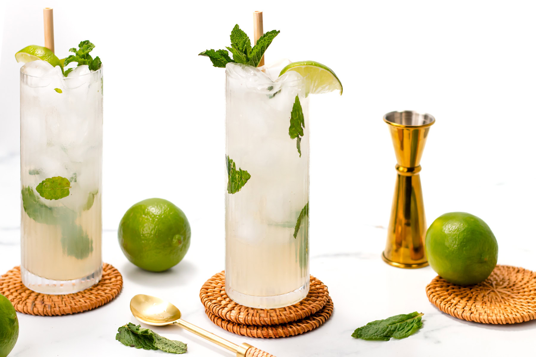 Two mojito cocktails with lime wedges and mint leaves on coasters, surrounded by whole limes, a gold jigger, and a spoon on a white surface.