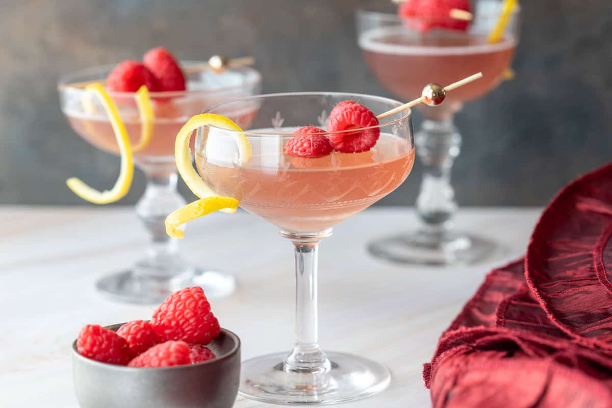 Three coupe glasses filled with a pink cocktail, each garnished with raspberries and a lemon twist. A small bowl of raspberries and a red cloth are next to the glasses.