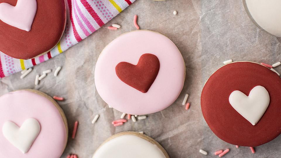 Round cookies decorated with heart-shaped icing in red, pink, and white, placed on parchment paper with scattered sprinkles and a striped cloth in the background.