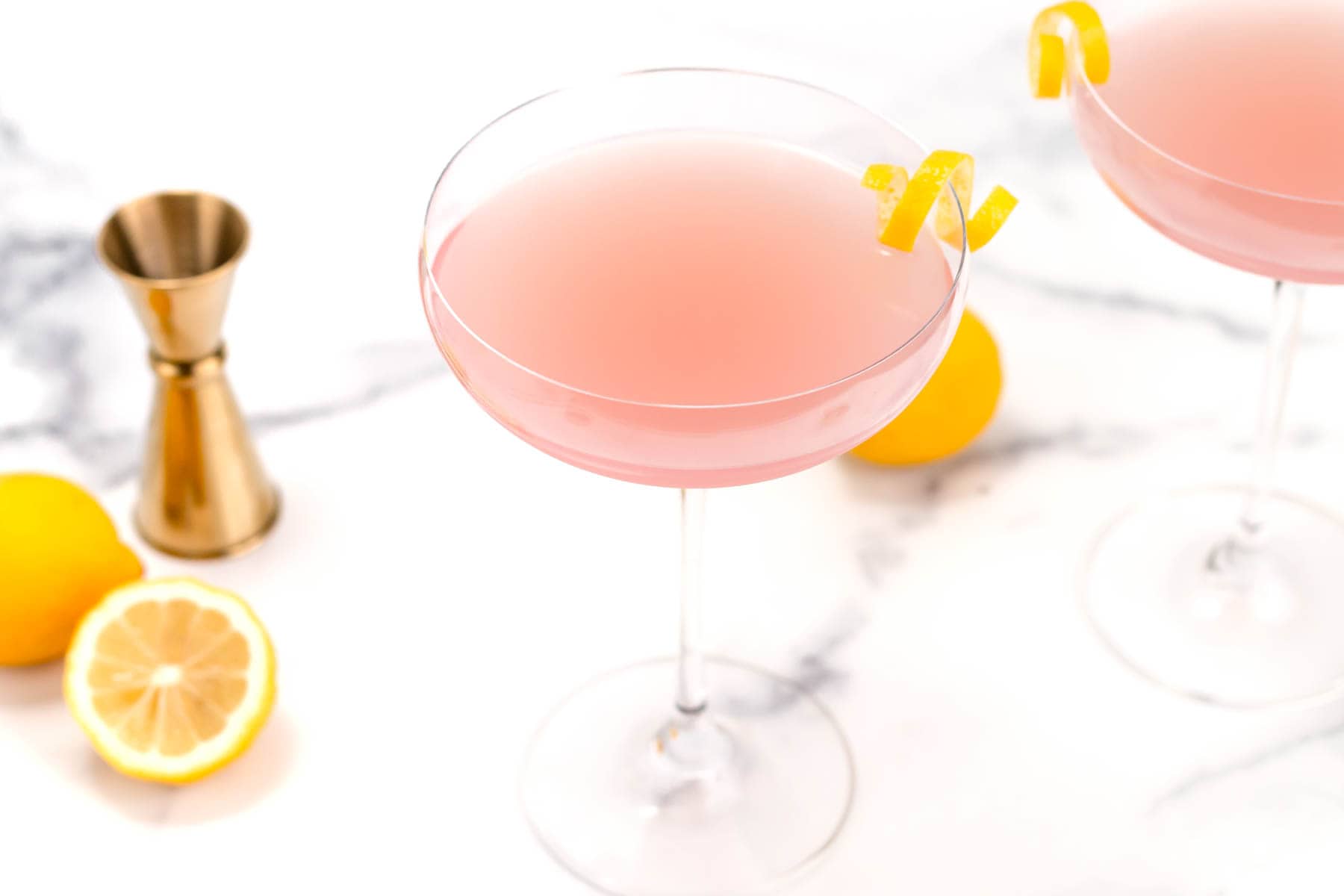 A pink cocktail in a coupe glass garnished with a lemon twist, with a jigger, whole lemons, and a lemon slice on a white marble surface.