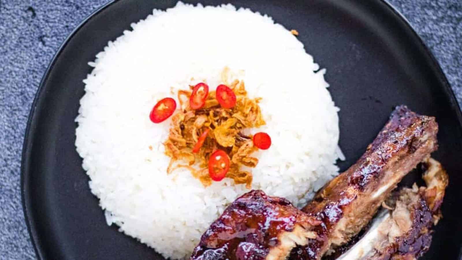 A black plate with white rice topped with fried shallots and sliced red chili, served alongside glazed ribs.