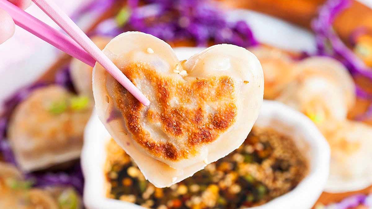 A pair of chopsticks holds a heart-shaped dumpling above a bowl of dipping sauce, with more dumplings and purple cabbage in the background.