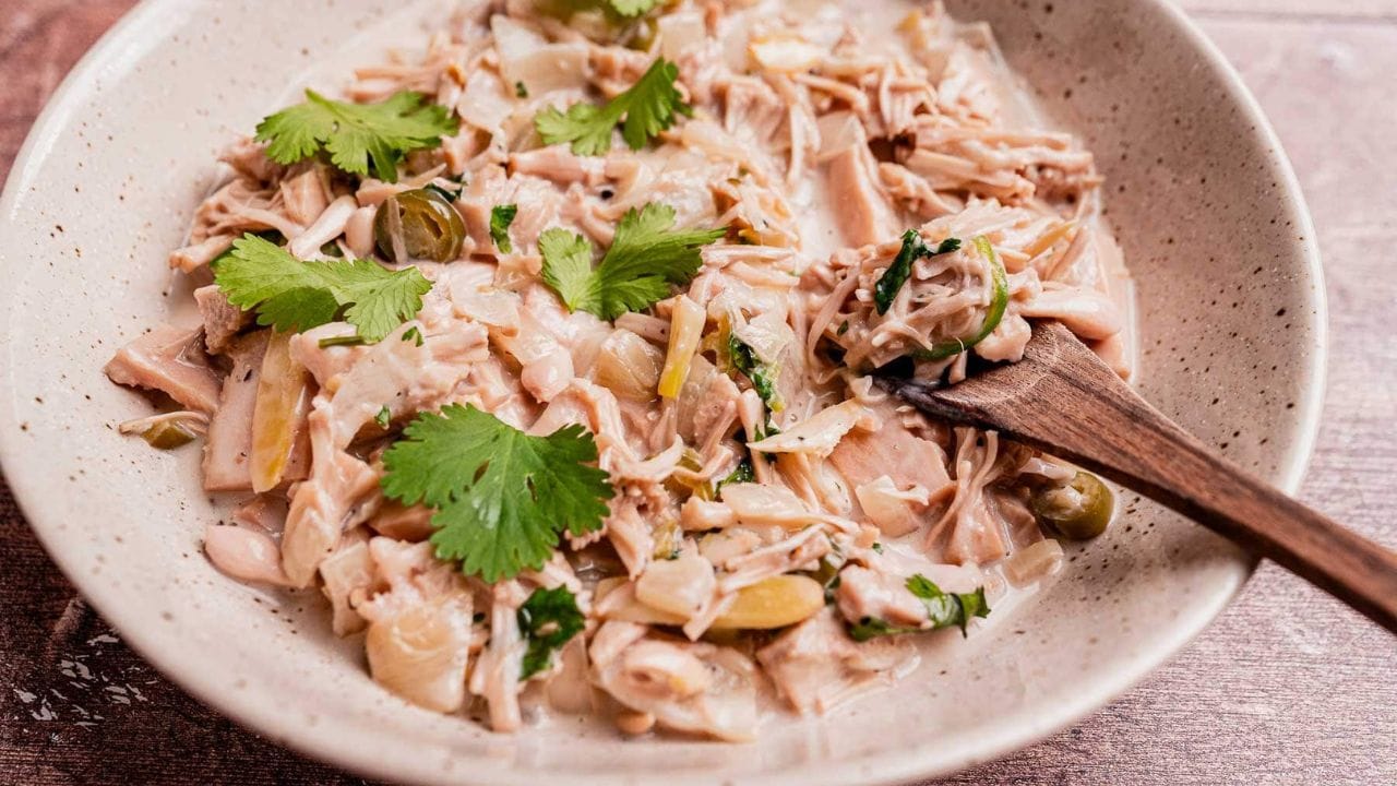 A bowl of shredded jackfruit stew garnished with fresh cilantro leaves, served with a wooden spoon.