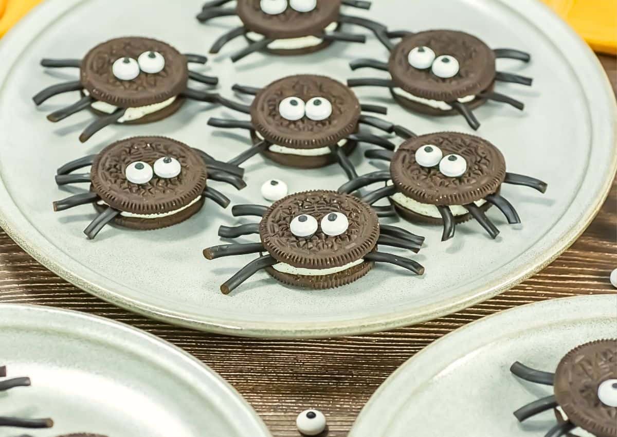 Chocolate sandwich cookies decorated with candy eyes and licorice pieces to resemble spiders, arranged on white plates.