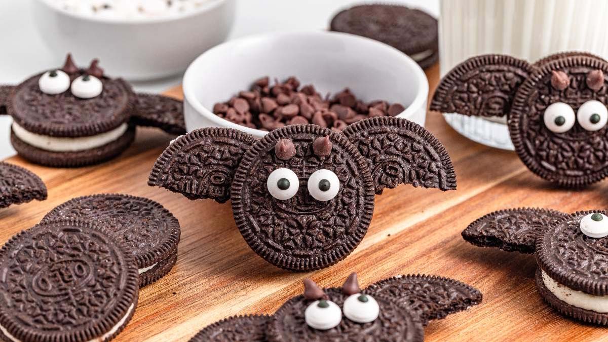 Chocolate sandwich cookies decorated with candy eyes and chocolate chips to look like bats, arranged on a wooden board with a bowl of chocolate chips and a glass of milk.