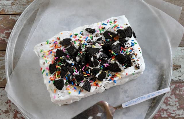 A rectangular ice cream cake topped with crushed chocolate cookies and colorful sprinkles sits on parchment paper with a cake server nearby.