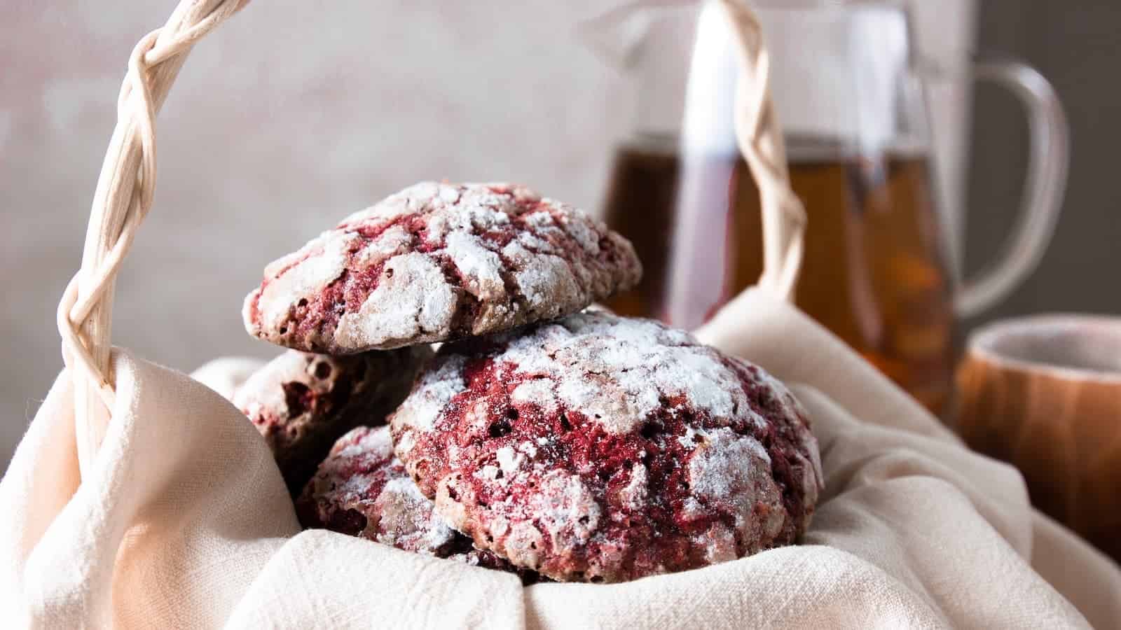 Three powdered red cookies rest on a beige cloth in a basket, with a glass pitcher and a cup in the blurred background.