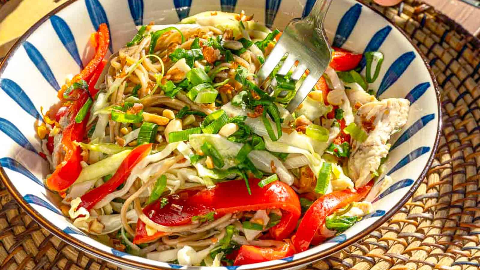 A bowl of noodle salad with sliced red bell peppers, cabbage, green onions, and crushed peanuts, with a fork resting inside.