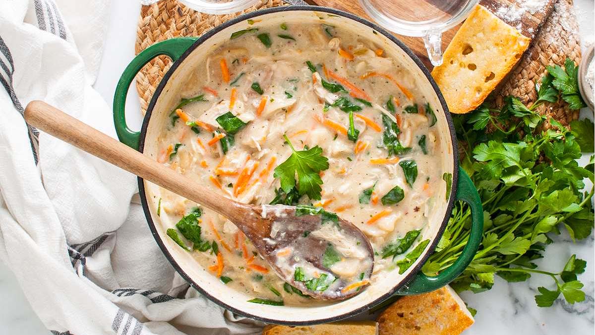 A pot of creamy soup with carrots, spinach, and herbs is stirred with a wooden spoon, surrounded by parsley, bread, and a cloth napkin.