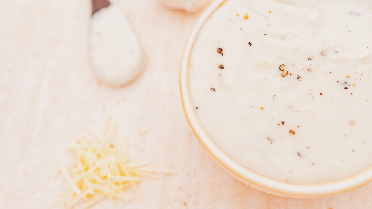 A bowl of creamy white sauce topped with black pepper, with shredded cheese and a spoon of sauce nearby on a light surface.