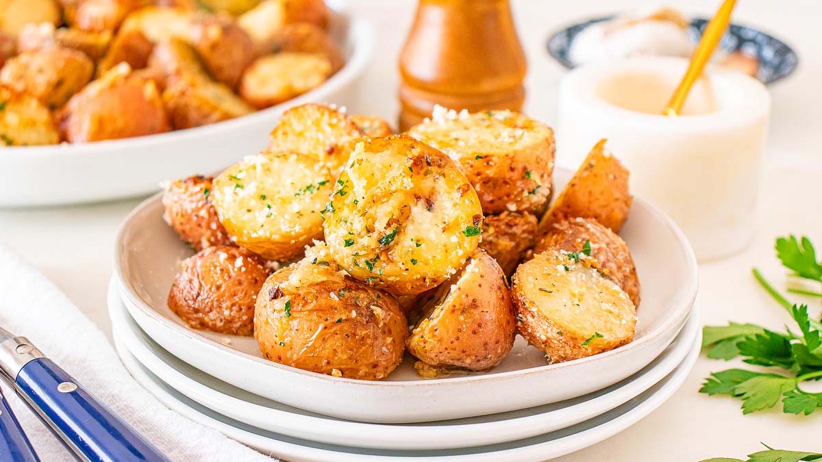 A plate of roasted potato halves garnished with herbs and grated cheese, with additional potatoes and condiments visible in the background.