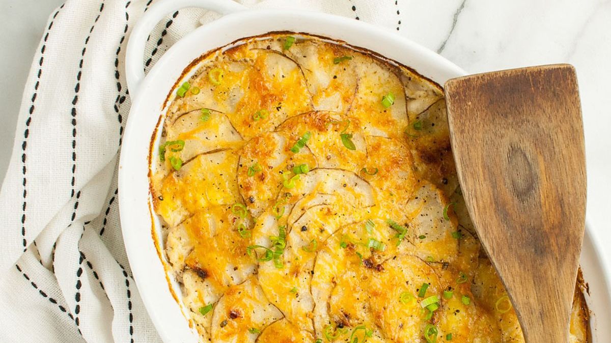 A baked dish of scalloped potatoes topped with melted cheese and chopped green onions sits on a white surface with a wooden spatula resting inside.