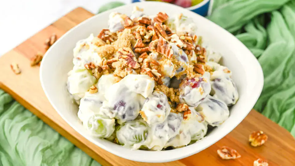 A bowl of grape salad topped with chopped pecans and brown sugar, placed on a wooden board with a green cloth underneath.