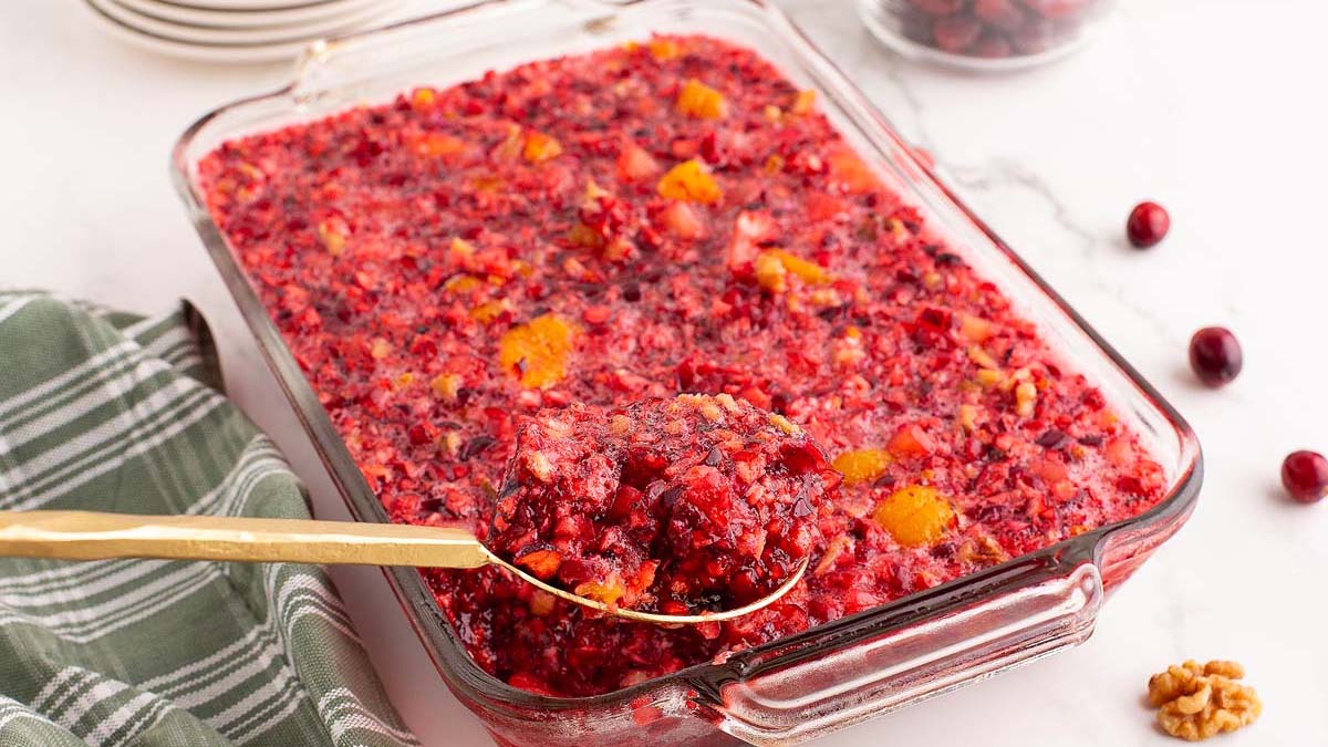 A glass baking dish filled with a red gelatin salad containing cranberries and orange pieces; a serving spoon holds a portion above the dish.
