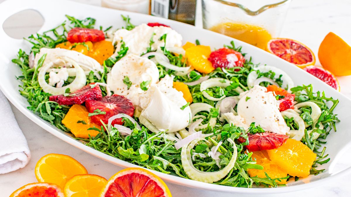 A white dish contains a salad with arugula, orange slices, red citrus segments, fennel, and dollops of soft cheese, garnished with herbs. A jar of dressing and citrus fruits are nearby.