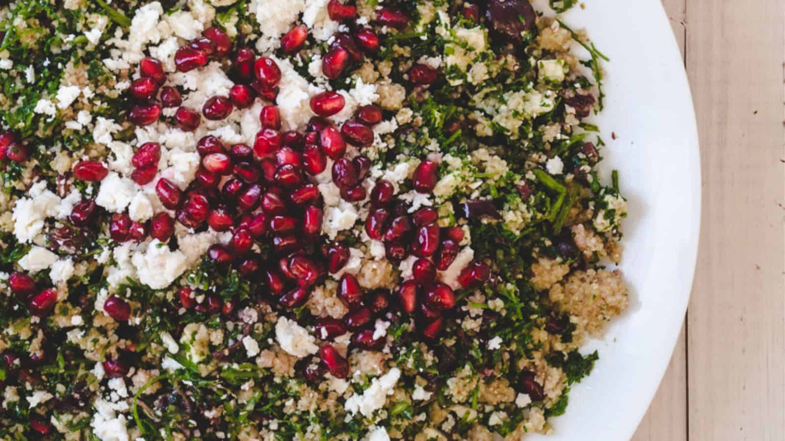 A plate of quinoa salad topped with chopped greens, crumbled feta cheese, and pomegranate seeds on a wooden surface.