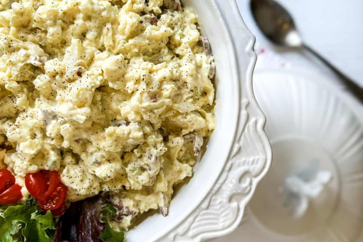 A bowl of creamy potato salad with herbs, black pepper, and a side of lettuce and cherry tomatoes, placed on a white plate next to a spoon.