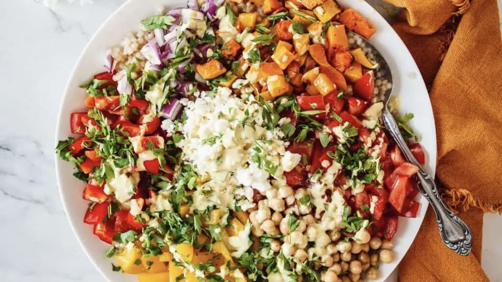 A bowl filled with chopped vegetables, chickpeas, couscous, feta cheese, and herbs, with a spoon resting inside.